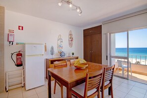 A dining area with a wooden table and chairs, a fire extinguisher, a refrigerator, and a view of the beach through a sliding glass door.