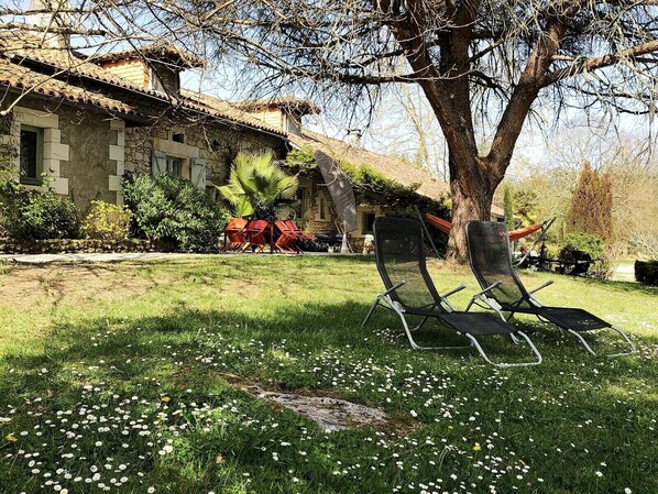 A stone building with a tiled roof, a tree, and two black lawn chairs in a grassy area.