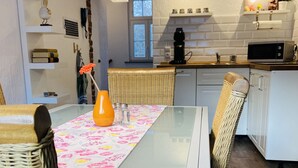A modern kitchen with white cabinets, a microwave, and a coffee machine. There is a dining table with a glass top and wicker chairs. - Gönn Dir Eine Auszeit - Raus ins Grüne - Komm in die Sötenicher Mühle (Kall)