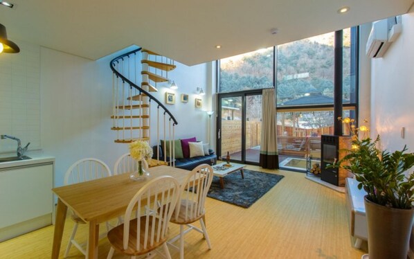 A modern kitchen with a wooden dining table and chairs, a staircase with a wooden railing, and a large sliding glass door leading to an outdoor area. - Wonju Cloud Pension (Wonju)