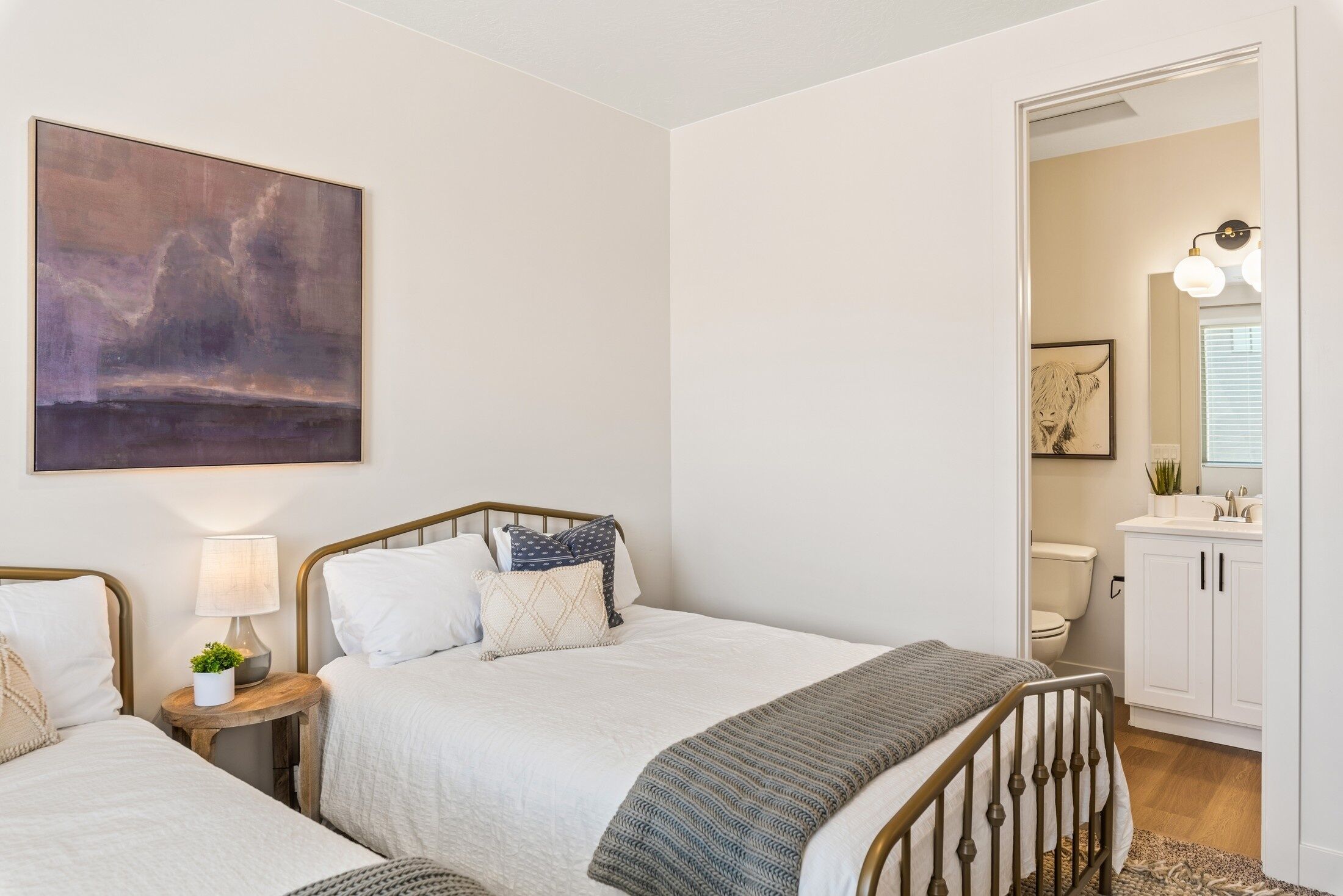 A bedroom with two beds, a bedside table with a lamp, a painting on the wall, and a bathroom visible through an open door.