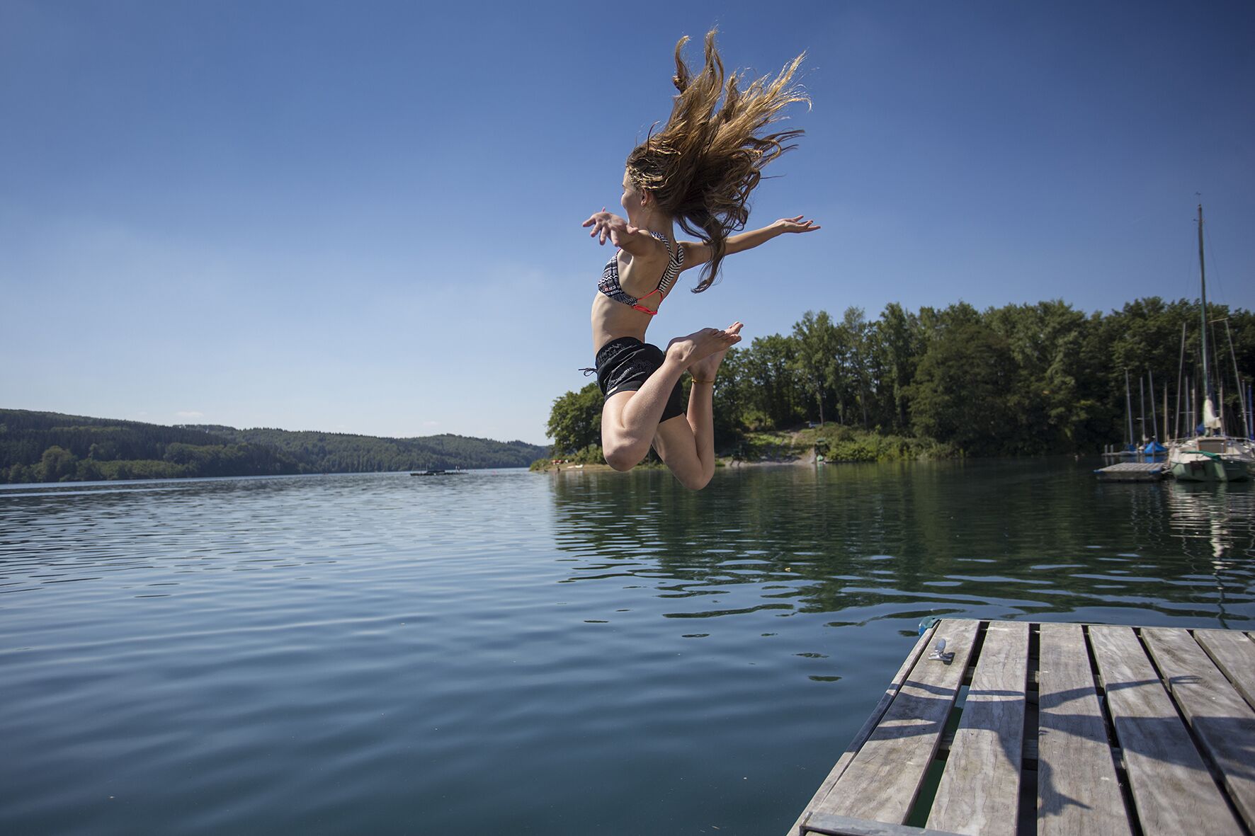 Une personne qui saute d’un quai dans un lac, avec des arbres et des bateaux en arrière-plan.