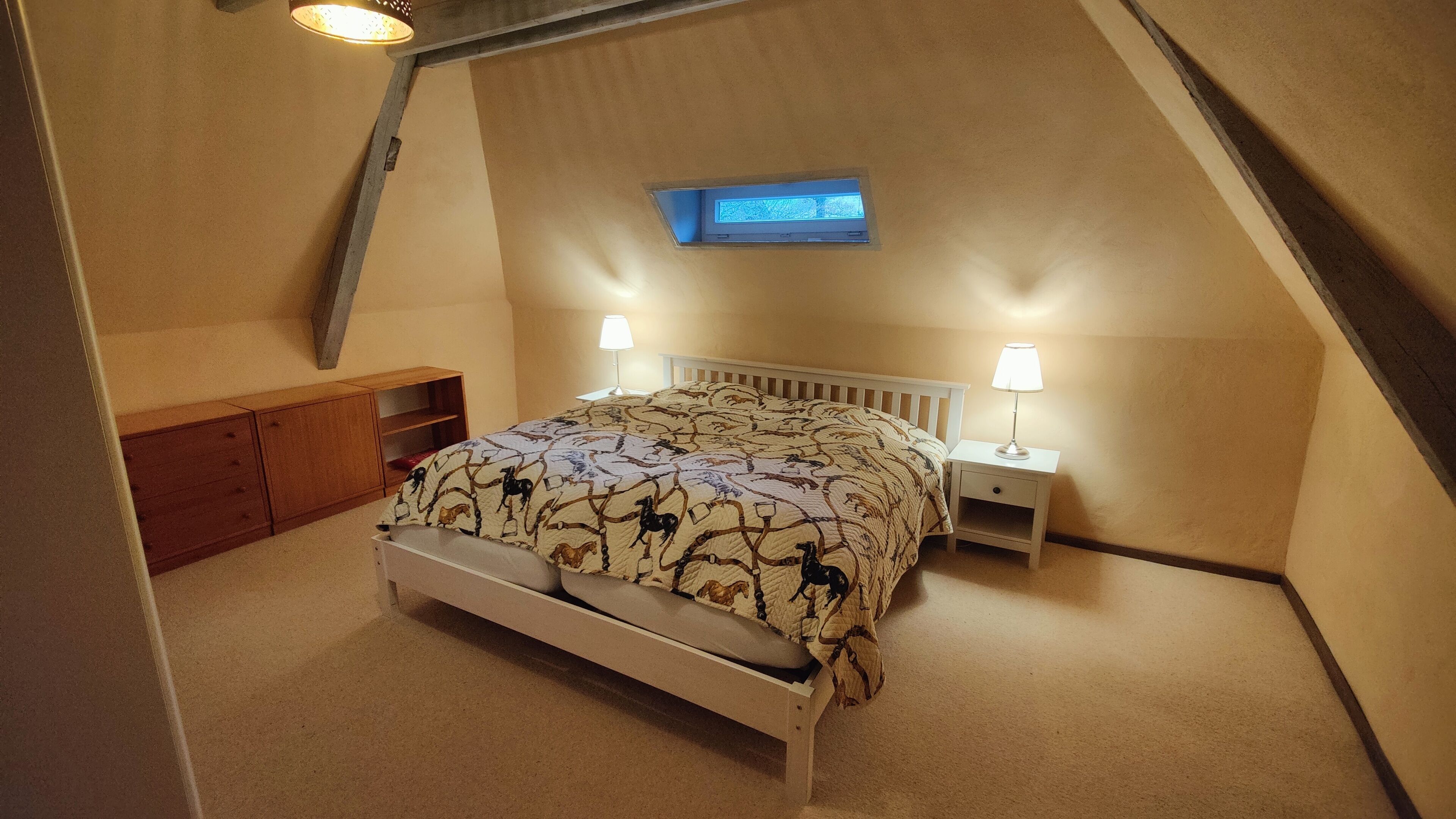 Une chambre avec un plafond en pente, un lit recouvert d’une couvre-lit à motifs, deux lampes de chevet, une petite étagère et un téléviseur fixé au mur.