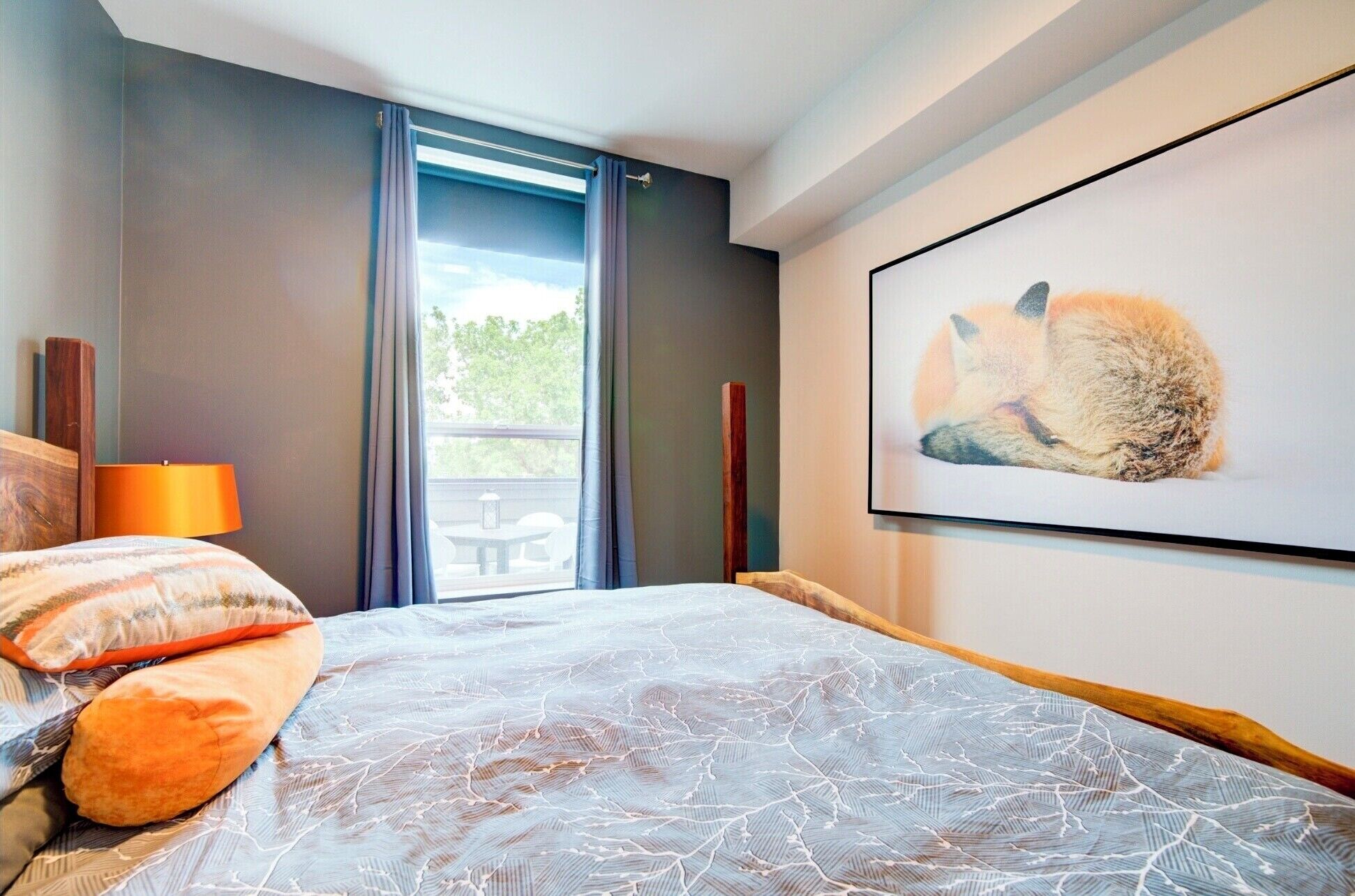 A bedroom with a bed, a window with curtains, a table and chairs outside, and a framed picture on the wall.