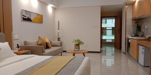 A modern hotel room with a bed, a sofa, a small table with a vase, and a kitchenette.