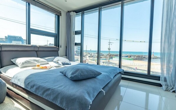 A modern hotel room with a large bed, a view of the beach, and a cityscape. - Jeju Judith Pension (Jeju City)