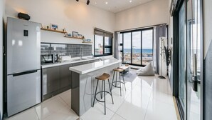 A modern kitchen with a large island, bar stools, and a view of the beach through the sliding glass door. - Jeju Judith Pension (Jeju City)
