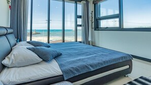 A modern bedroom with a large bed, a view of the beach, and a cityscape through large windows. - Jeju Judith Pension (Jeju City)