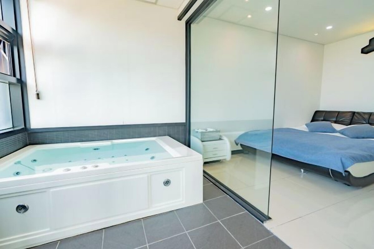 Quarto moderno com box de vidro, uma banheira grande e branca, cama com roupa de cama azul e cabeceira escura.