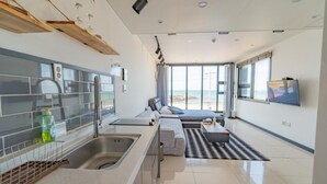 A modern hotel room with a kitchen, a sofa bed, a TV, and a view of the beach. - Jeju Judith Pension (Jeju City)