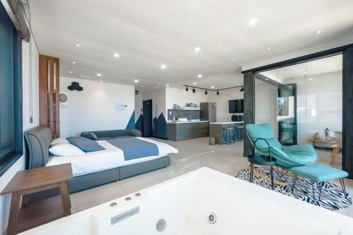 Quarto de hotel moderno com uma cama grande, área de estar com uma poltrona, uma mesa pequena, banheira e cozinha ao fundo.
