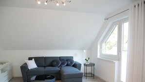 A modern living room with a grey sectional sofa, a small round coffee table, and a grey ottoman. There is a side table with a vase of flowers, a lamp, and a window with curtains. - Fewo Wagner +nur 10 Gehminuten bis zum Ostseestrand+ - Ferienwohnung Wagner (Koserow)