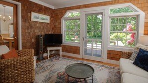 A living room with a television, wicker furniture, a patterned rug, and a view of a garden through large windows. - Family Fun, Spacious+Game Room (Oak Bluffs)