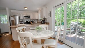 A kitchen with white cabinets, a central island with chairs, a dining table with a floral tablecloth, and a large window overlooking a deck. - Family Fun, Spacious+Game Room (Oak Bluffs)