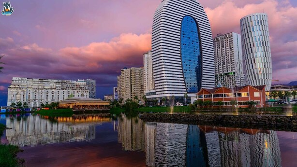 Un paisaje urbano con modernos rascacielos, una masa de agua que refleja las construcciones, y un cielo despejado.