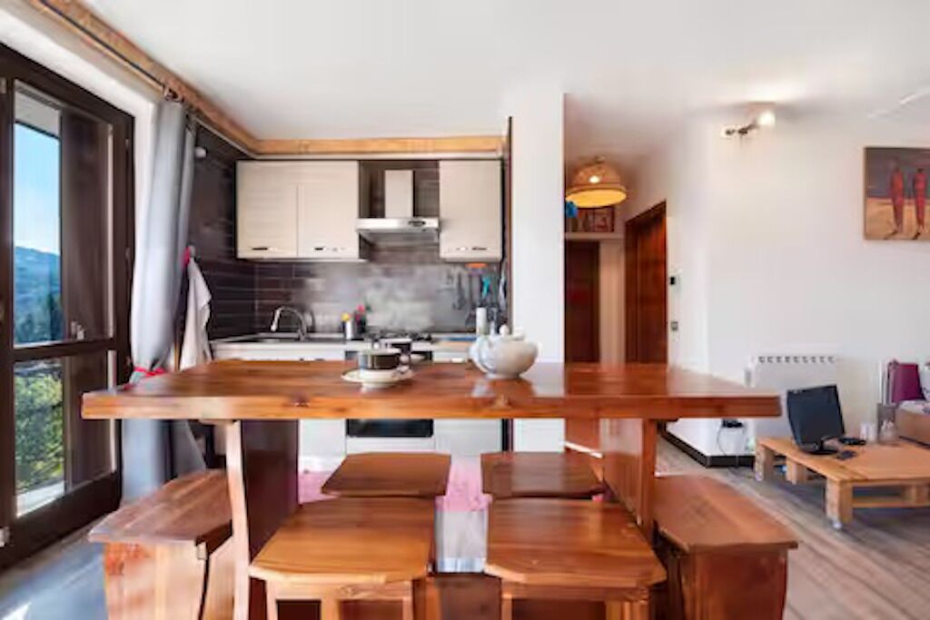 Une cuisine moderne avec des plans de travail et des meubles en bois, un coin repas avec une grande table et des bancs en bois, et une vue sur l’extérieur grâce à une grande fenêtre.