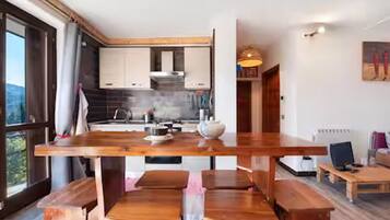 Une cuisine moderne avec des plans de travail et des meubles en bois, un coin repas avec une grande table et des bancs en bois, et une vue sur l’extérieur grâce à une grande fenêtre.