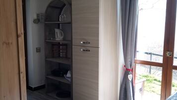 A room with a wooden wardrobe, a shelf unit with various items, a window with curtains, and a door leading outside.