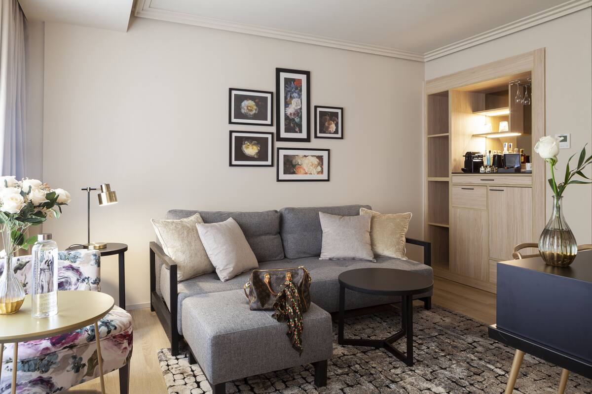 Een moderne woonkamer met een grijze bank, een ronde salontafel en een eettafel met stoelen. Er hangen ingelijste foto's aan de muur, een vaas met bloemen en er is een keuken te zien door een open deur.