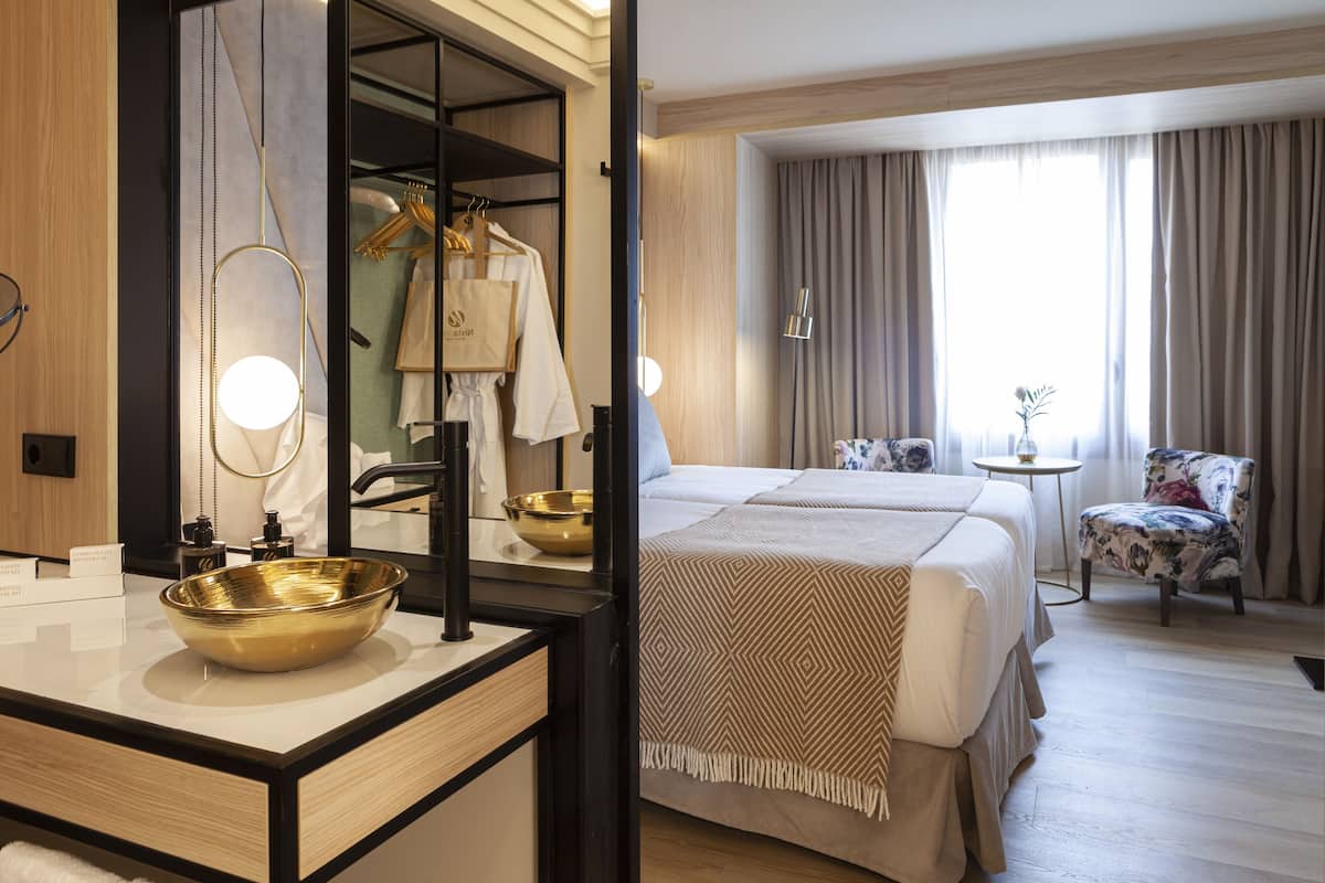 Hotelkamer met een grote spiegel, een gouden schaal op de wastafel, een bed met een geborduurde deken, een klein rond tafeltje en een fauteuil met bloemenprint.