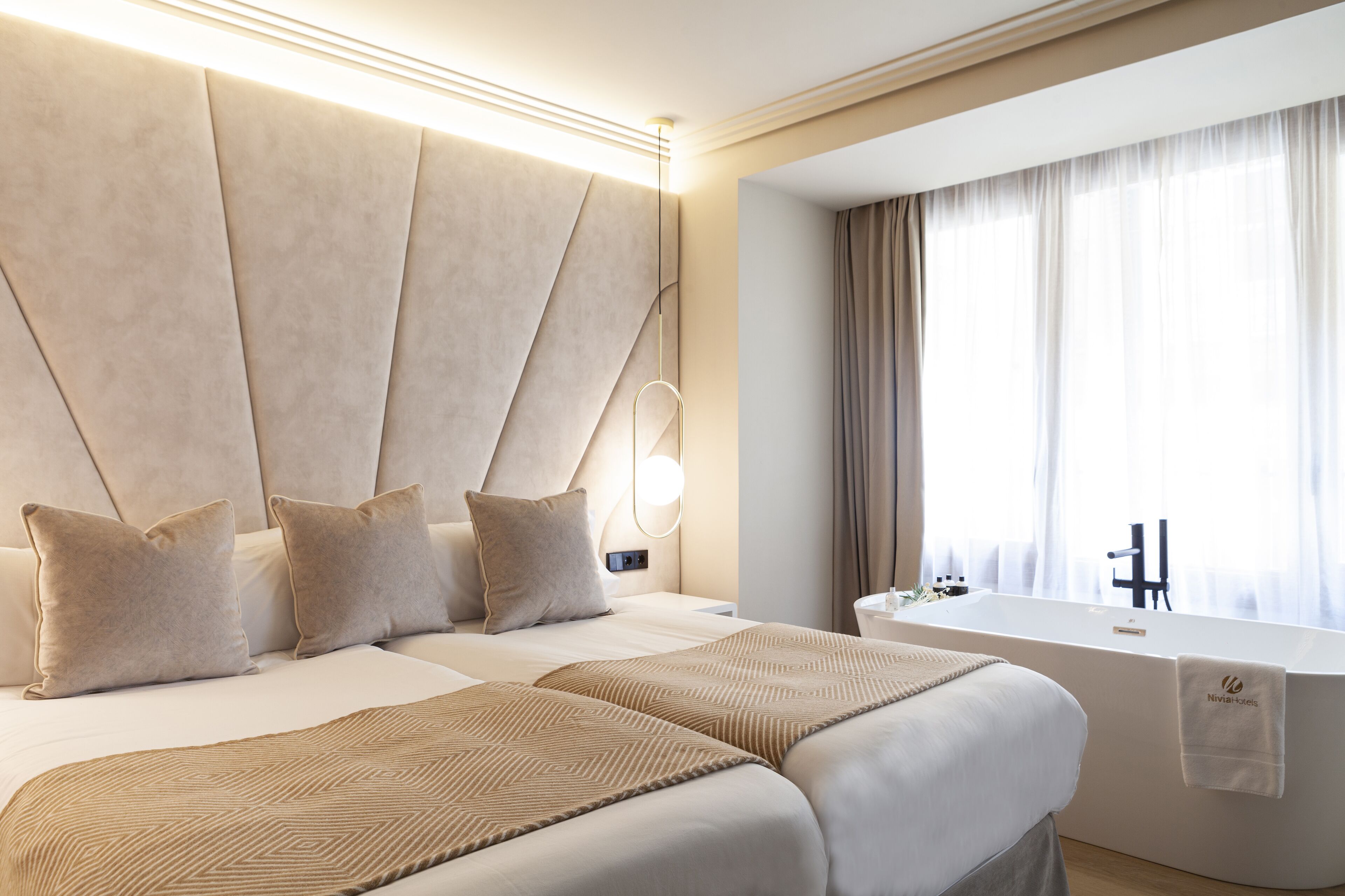 Una habitación de hotel moderna con una cama grande, una zona de estar separada, una bañera y un amplio ventanal con cortinas.