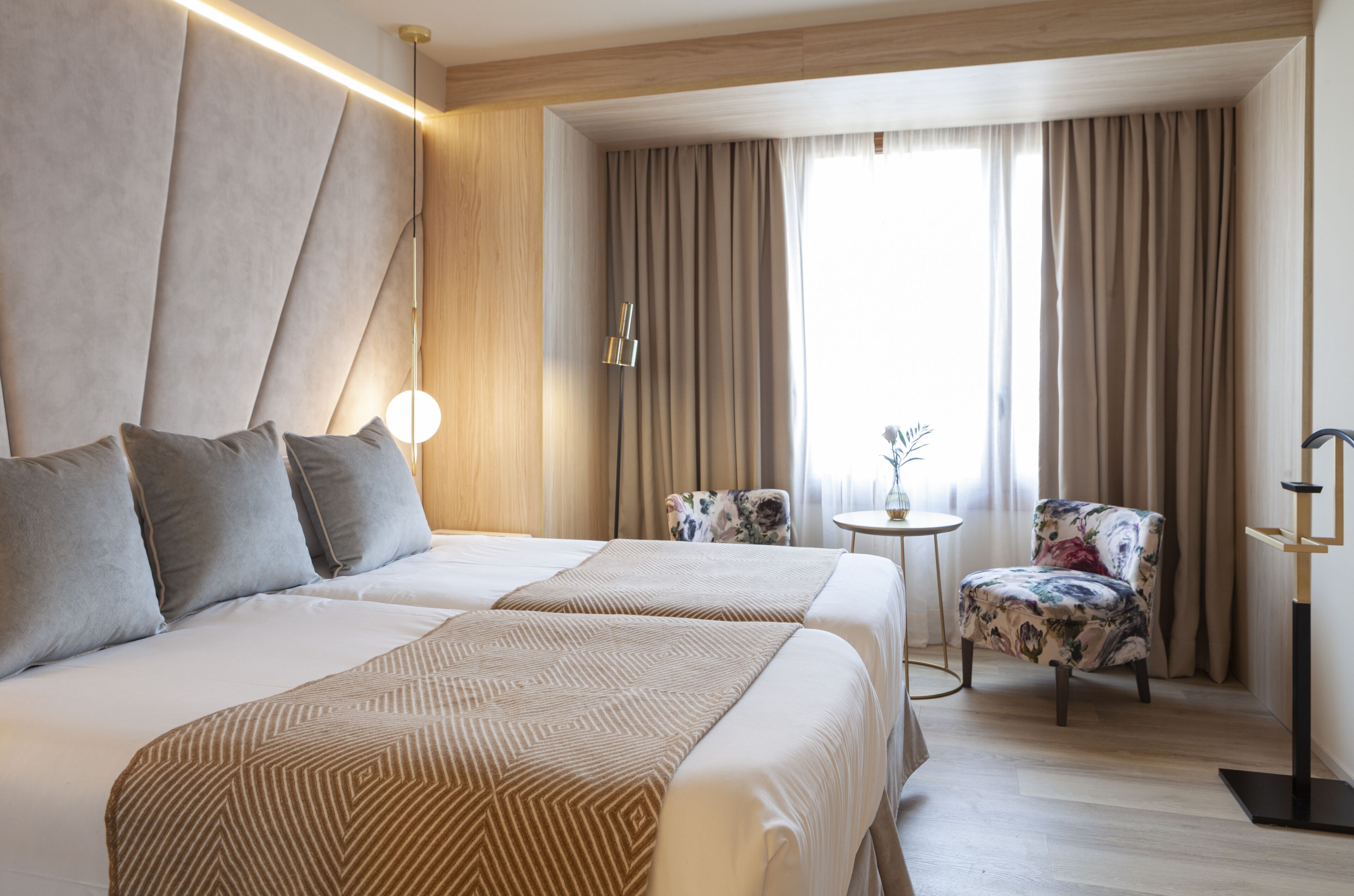 Een hotelkamer met twee bedden, een klein rond tafeltje, een stoel en een groot raam met gordijnen.