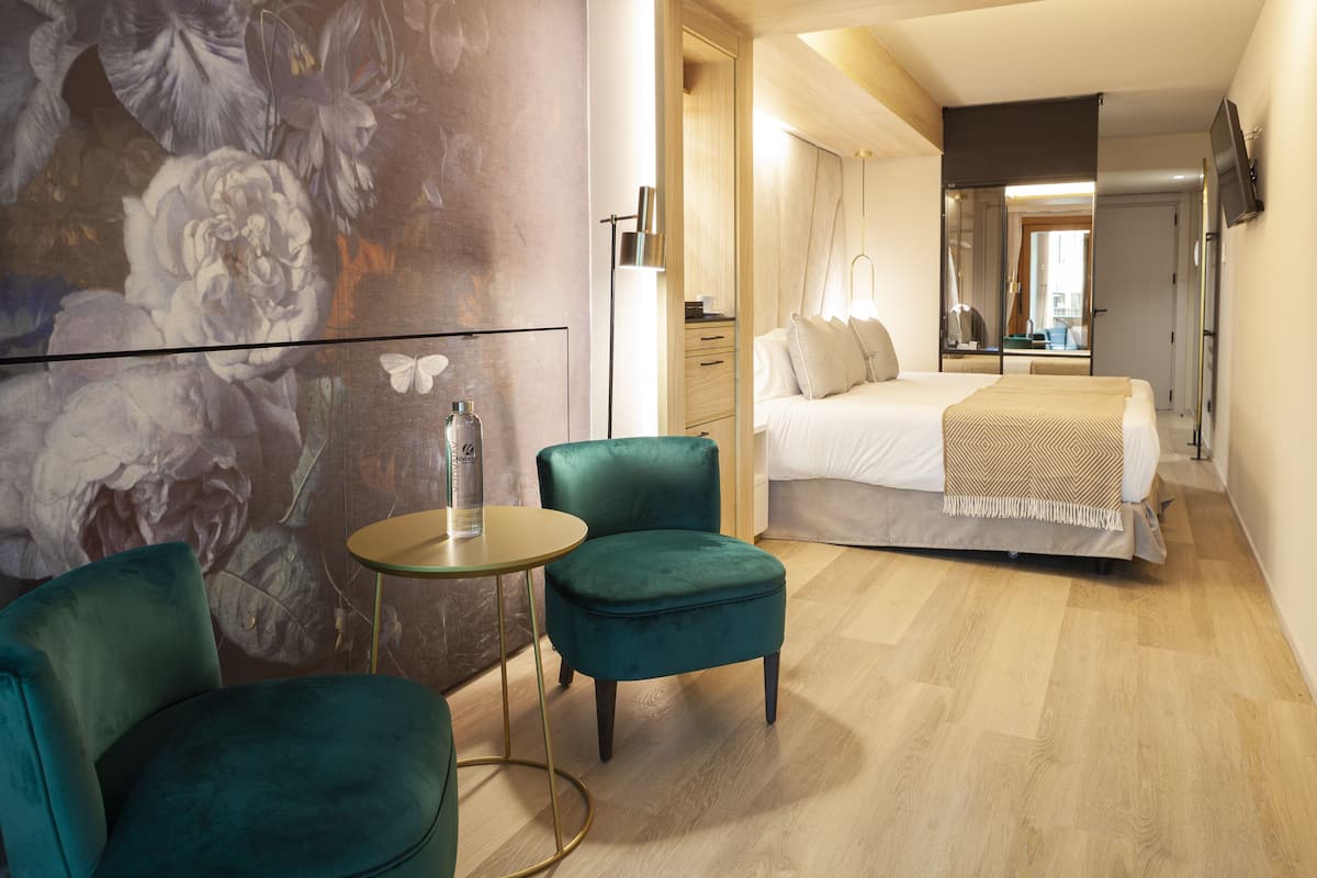 Een moderne hotelkamer met een groot bed, een groene fauteuil, een klein rond tafeltje en een bloemenmuur.