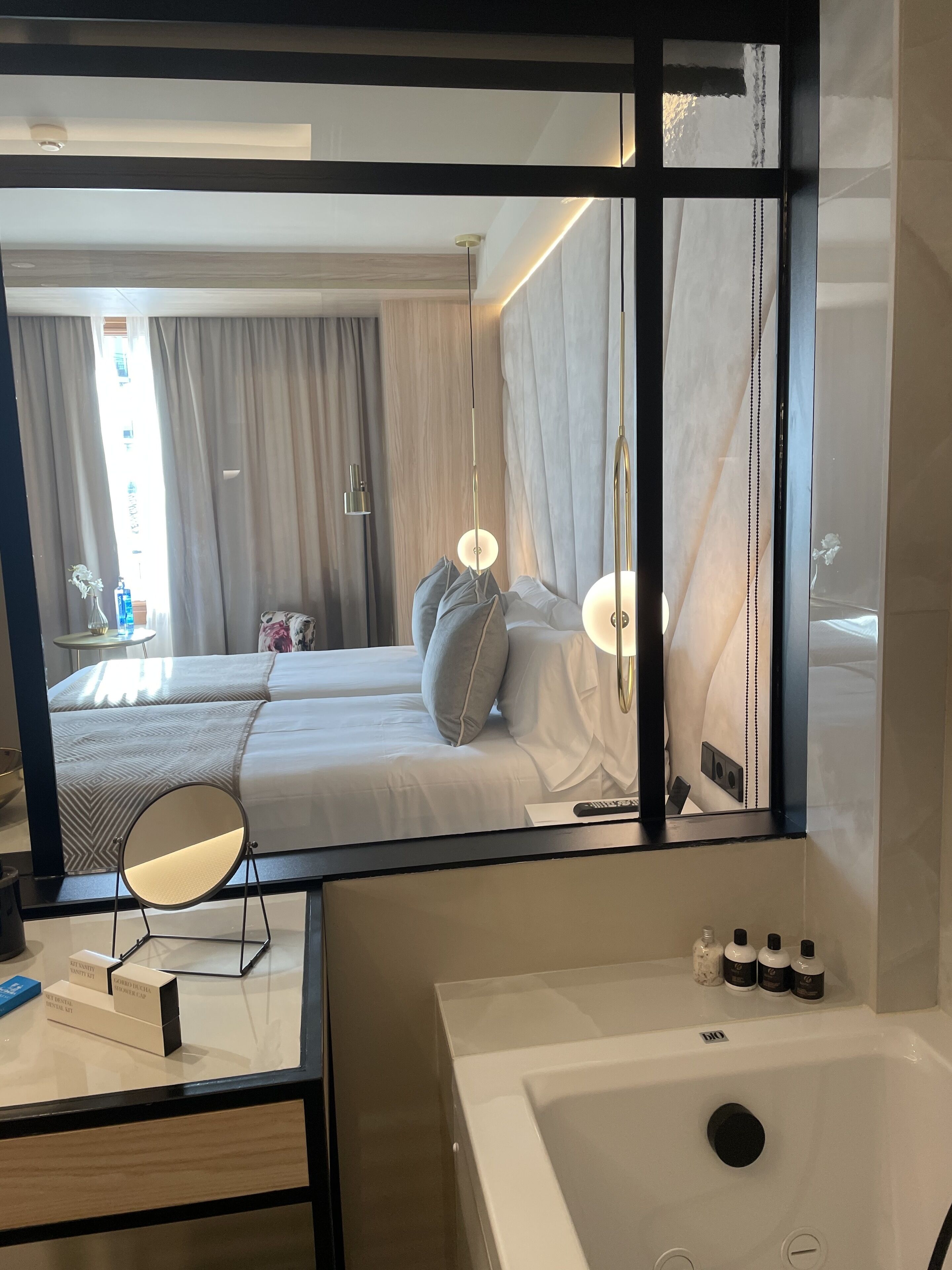 Habitación de hotel con un espejo grande que refleja la cama, una mesita de noche y un baño con bañera y artículos de aseo.
