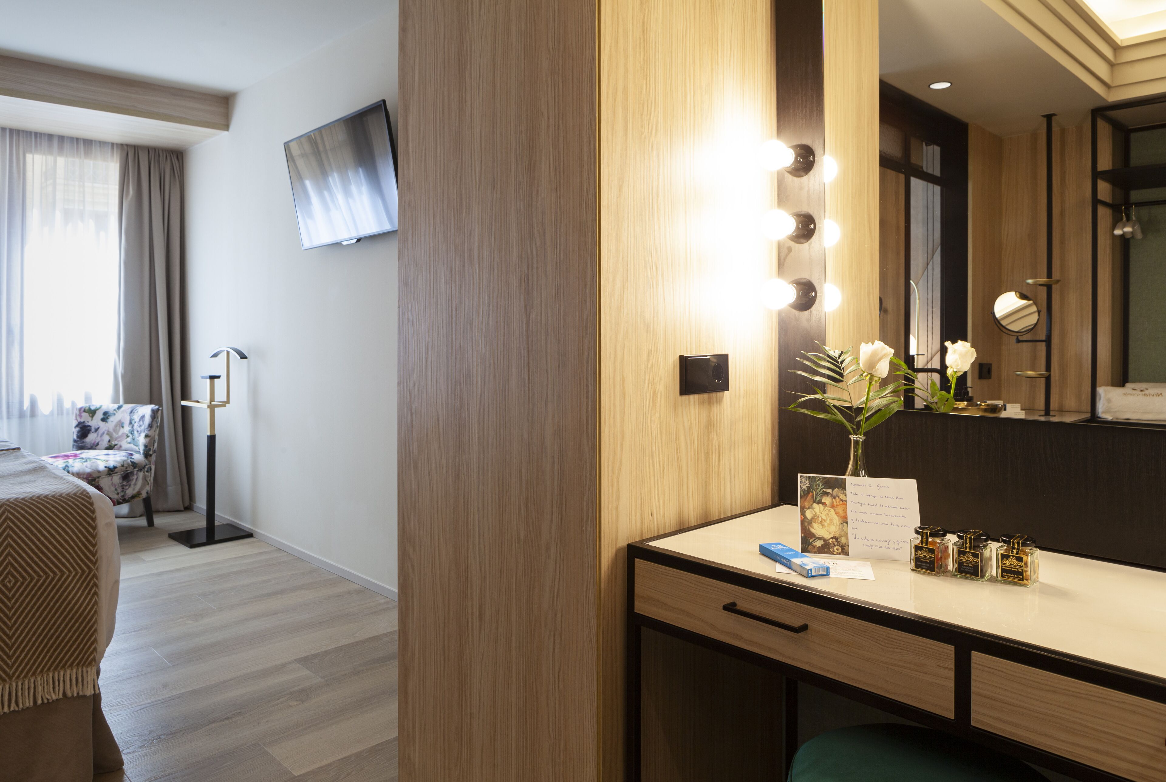 A hotel room with a wooden desk, a mirror with lights, a chair, a TV, and a window with curtains.
