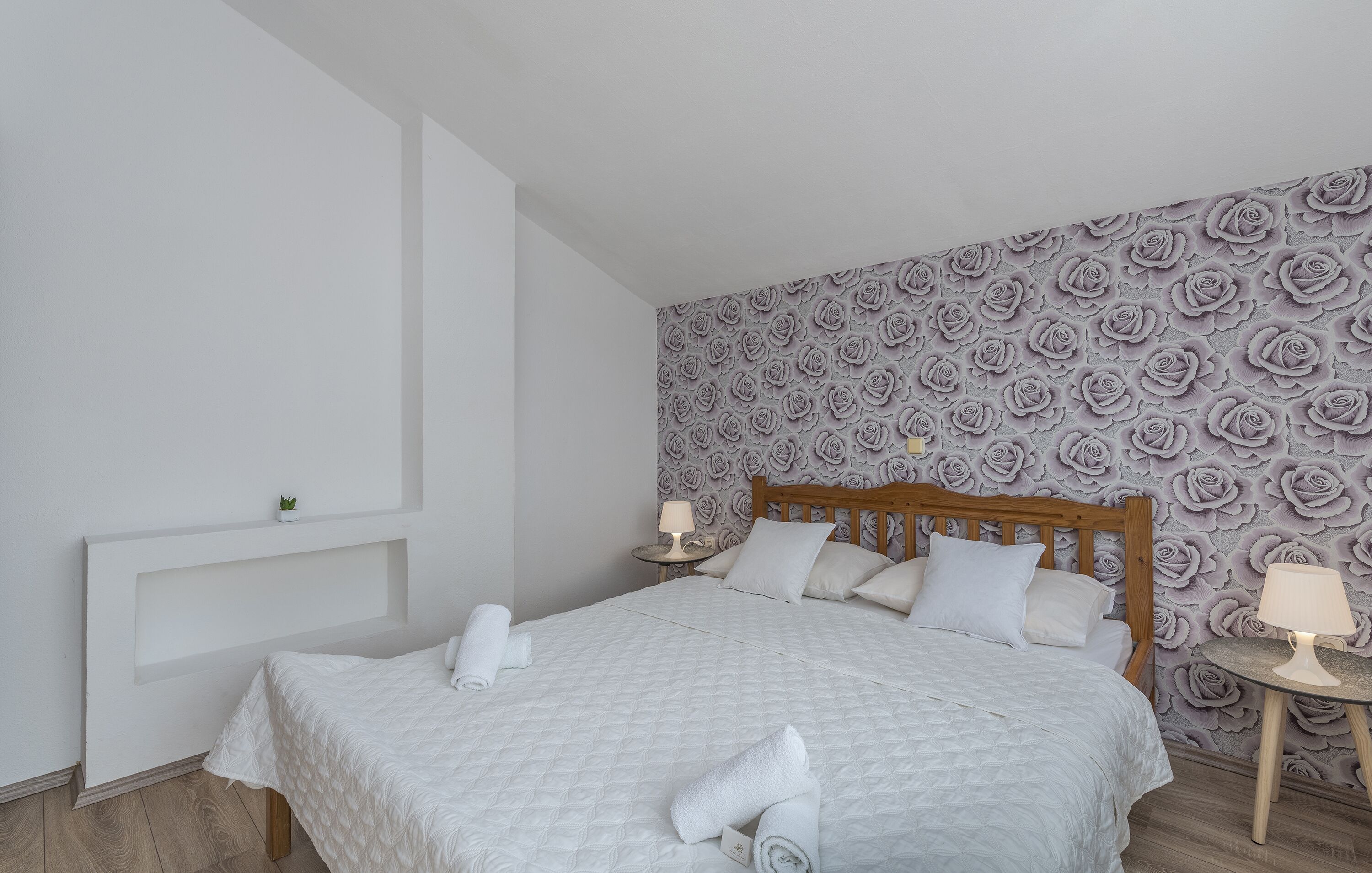 A neatly made bed with a white quilt, a wooden headboard, and a wall with a floral pattern.