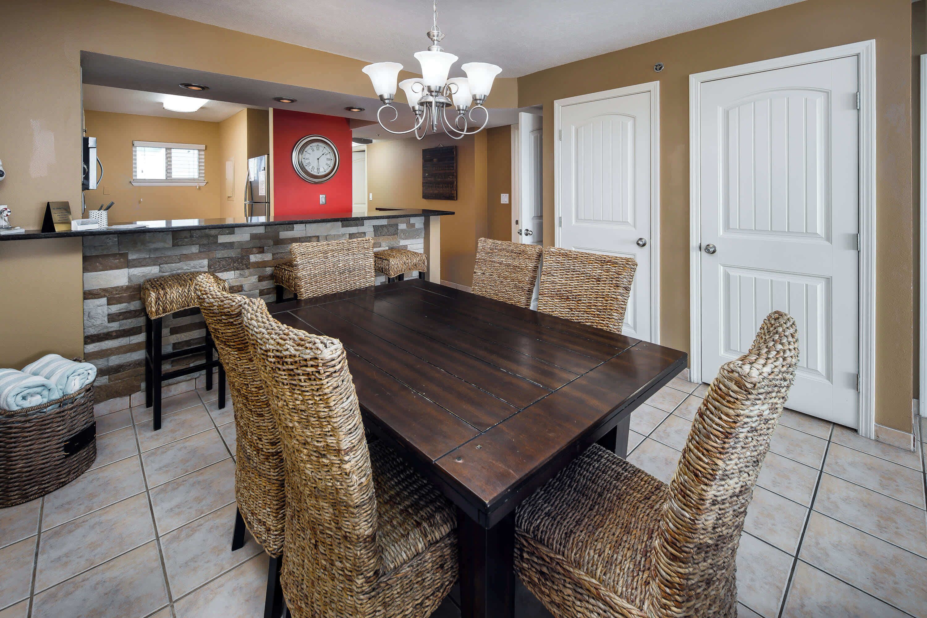 A dining area with a dark wooden table and woven chairs, a kitchen in the background, and a red-framed clock on the wall.