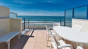 Ein Balkon mit weißem Tisch und Stühlen, der Blick auf einen Strand mit Meer und blauem Himmel bietet.