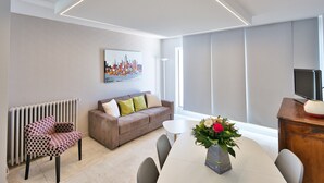 Ein modernes Wohnzimmer mit einer Couch, einem Esstisch mit Stühlen, einer Heizung, einem Bild an der Wand und einem Fernseher auf einem Holzbodenständer.