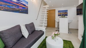 Uma sala de estar moderna com um sofá cinzento, uma mesa de centro branca com flores, uma escada e uma pintura de paisagem urbana.