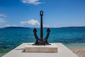 Une sculpture d’ancre noire, posée sur une plateforme en béton, au bord d’une mer d’un bleu limpide, avec une terre lointaine à l’horizon.