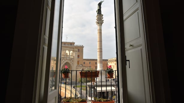 A view from a balcony with a railing, overlooking a plaza with a tall column and a building with arches.