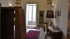 A hotel room with a wooden desk, a chair, a lamp, a coffee maker, and a water bottle on the desk. There are two patterned rugs on the floor.