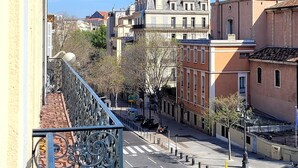 Ein Balkon mit aufwendiger Metallgliederung und Fliesenboden, der auf eine Stadtstraße mit Gebäuden und blauem Himmel blickt.