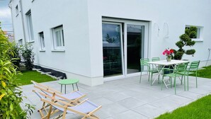 Un patio con mesa y sillas, una silla lounge de madera y un pequeño jardín.
