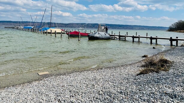 Una playa de guijarros con barcos amarrados a un muelle de madera que se adentra en aguas tranquilas.