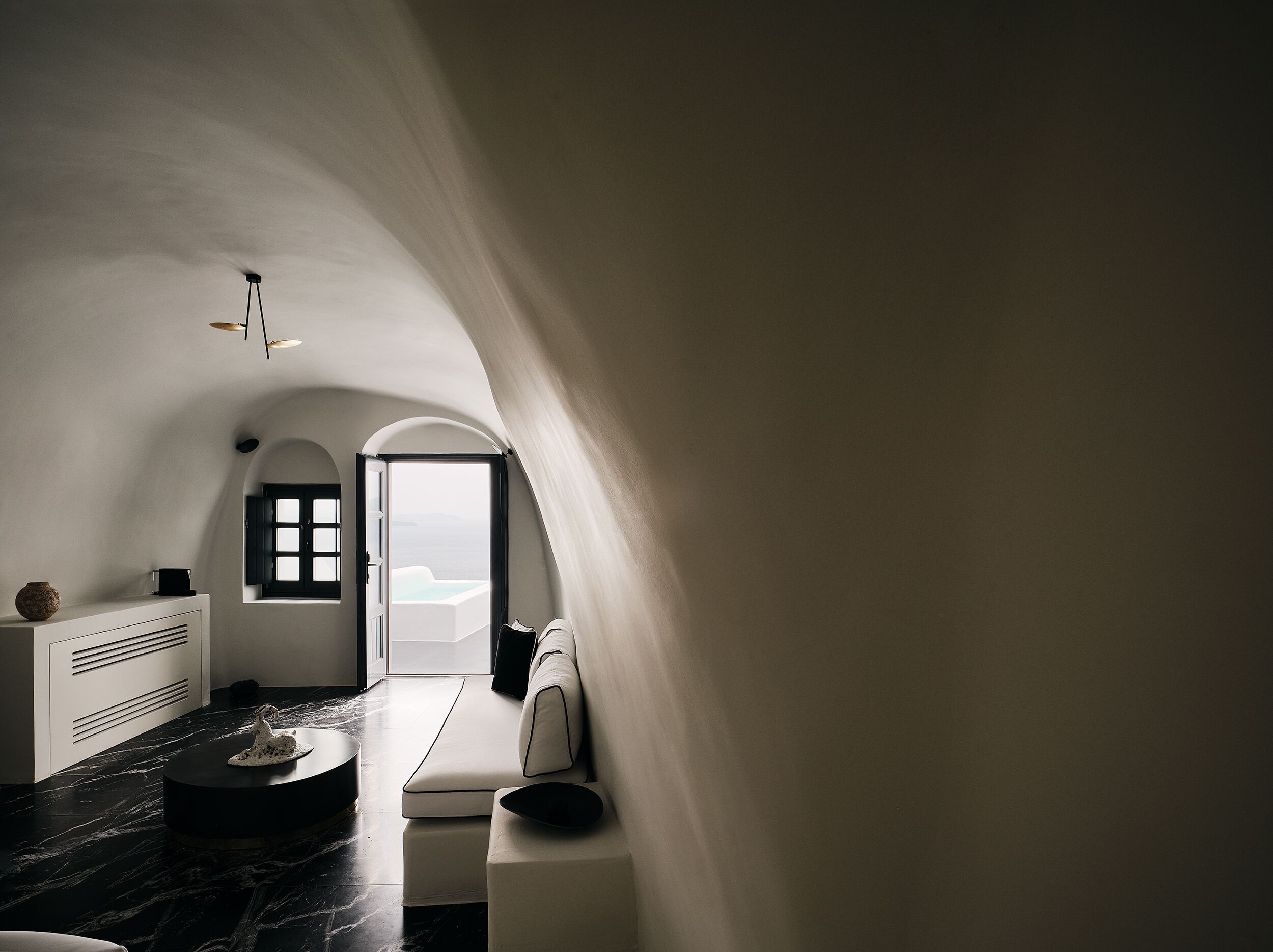 Un intérieur moderne avec un plafond incurvé, un canapé aux lignes épurées, une table ronde et une vue sur la mer, visible à travers une porte ouverte.
