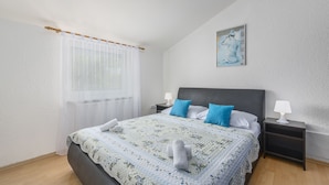 A bedroom with a bed, two nightstands, a window with sheer curtains, a painting on the wall, and a white lamp on one of the nightstands.