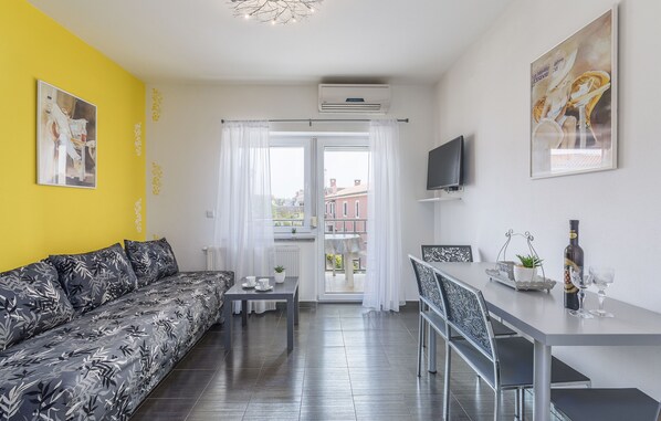 A modern living room with a sofa, dining table, and chairs. There is a TV mounted on the wall, a window with curtains, and a balcony visible through the window. - Villa Maja 4 Beachfront Apartment (Funtana)