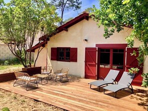 Une terrasse avec des meubles d’extérieur, une grande plante en pot et un bâtiment aux volets rouges.
