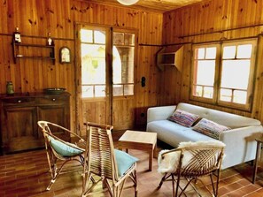 Interior acogedor de una cabaña de madera con un sofá, dos sillones de mimbre, una mesita y un ventanal con vistas.