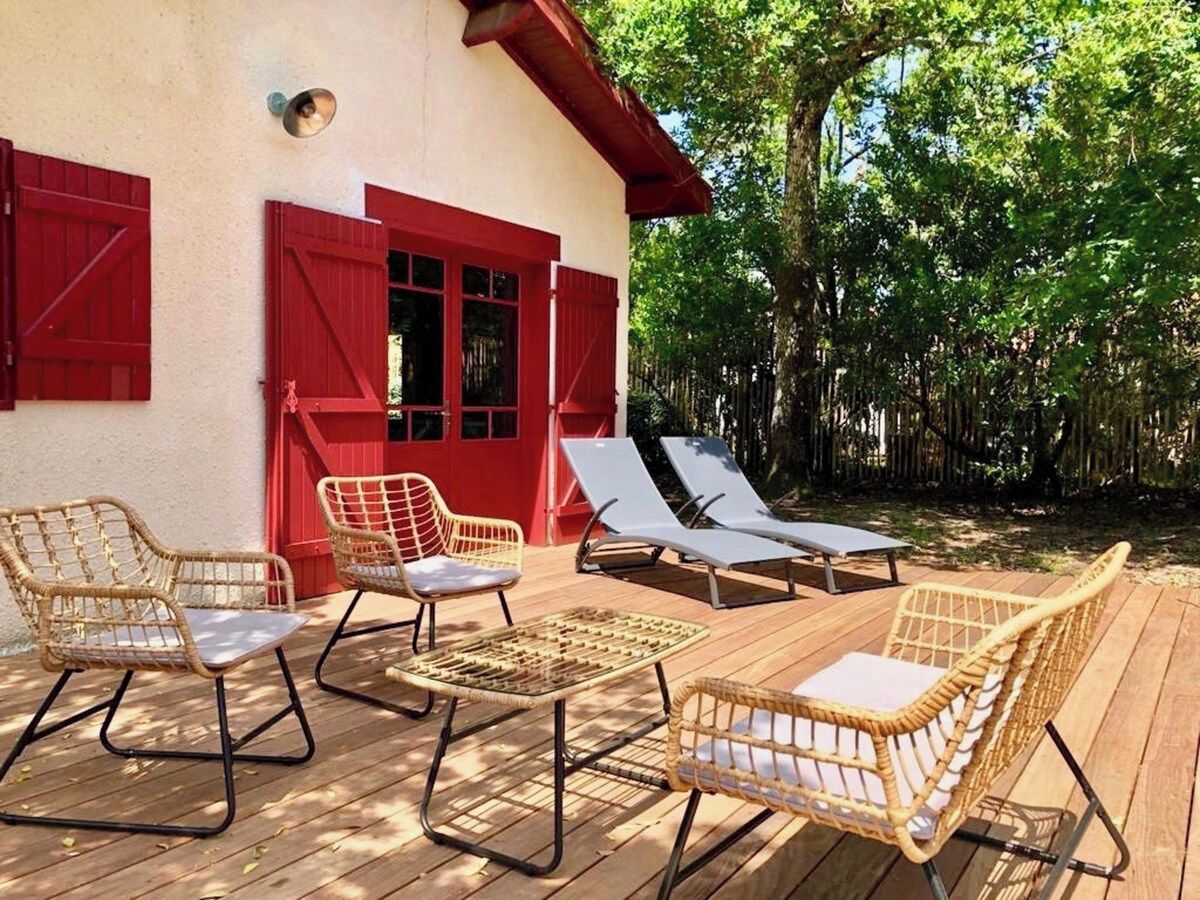 Une terrasse en bois avec des meubles en osier, notamment des chaises et une table, et un bâtiment rouge avec des volets.