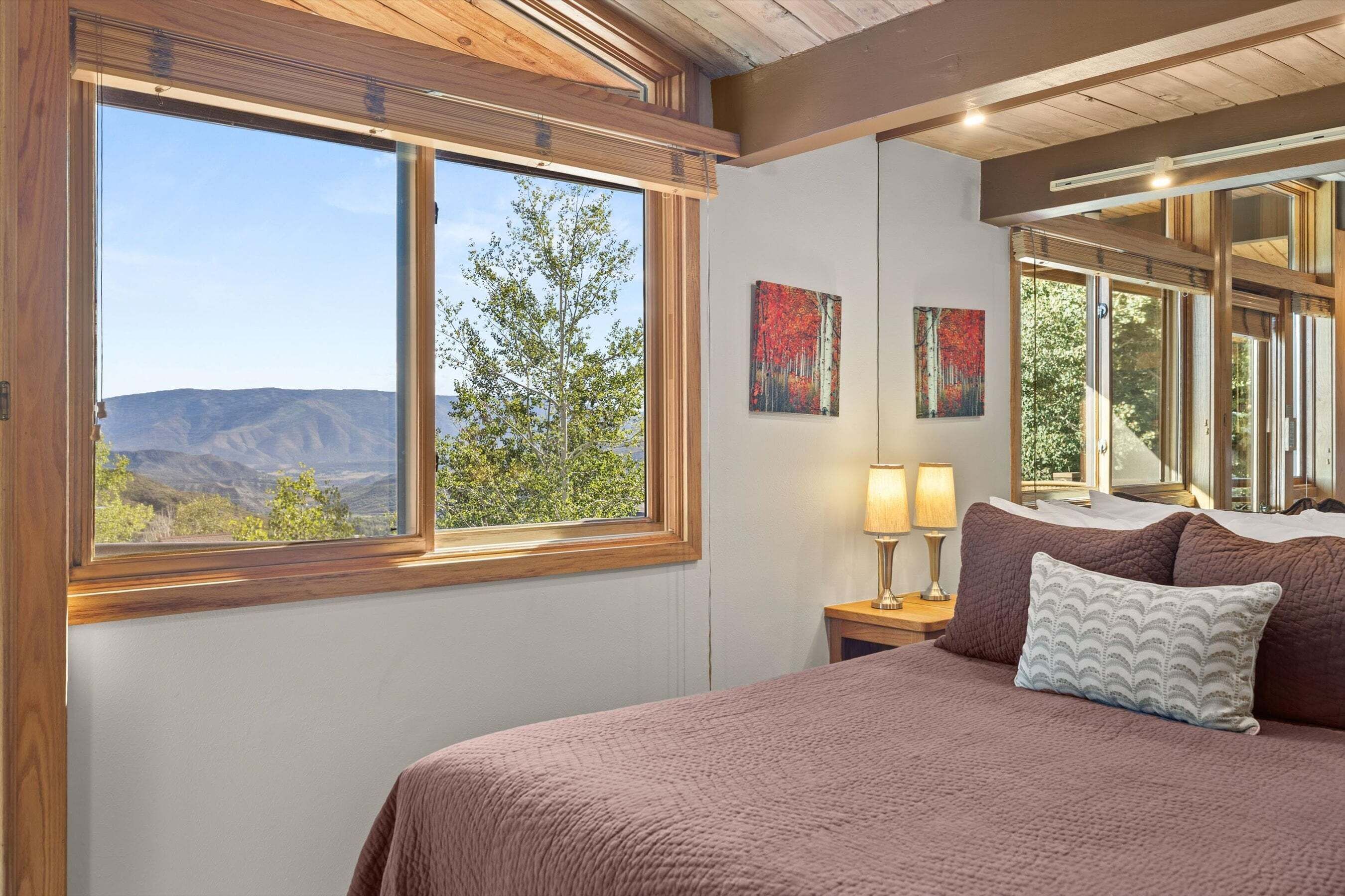 A bedroom with a large window, a bed with a pink blanket, two bedside lamps, and two framed paintings.