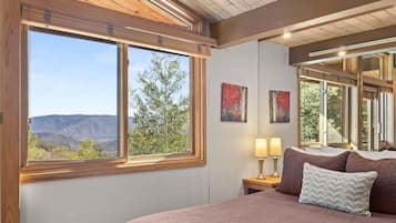 A bedroom with a large window, a bed with a pink blanket, two bedside lamps, and two framed paintings.