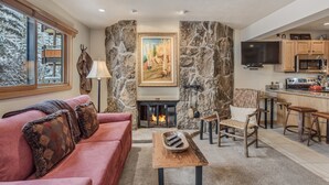 A cozy living room with a fireplace, stone walls, a red sofa, and a kitchen area with wooden cabinets and a flat-screen TV. - 🏔️Ski Access~Hot Tub~Studio Premier 305🏔️ (Snowmass Village)