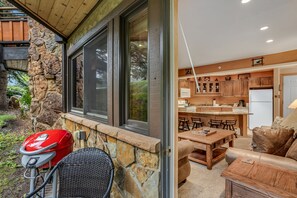 Eine Mauer aus Naturstein mit einem großen Fenster, ein Grill und ein Korbstuhl. Innen befindet sich eine Küche mit Holzausstattung, ein Essbereich mit Tisch und Stühlen sowie eine Ledersitzgruppe.